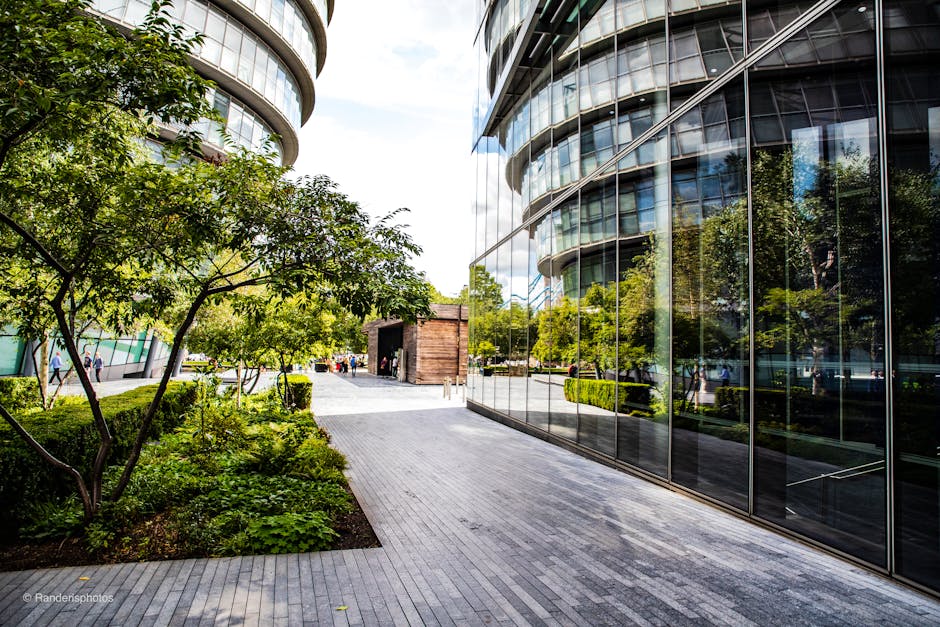 A modern urban garden situated alongside a sleek, curved glass building reflecting surrounding trees and sky. The garden features a landscaped section with lush green foliage, small trees with texture