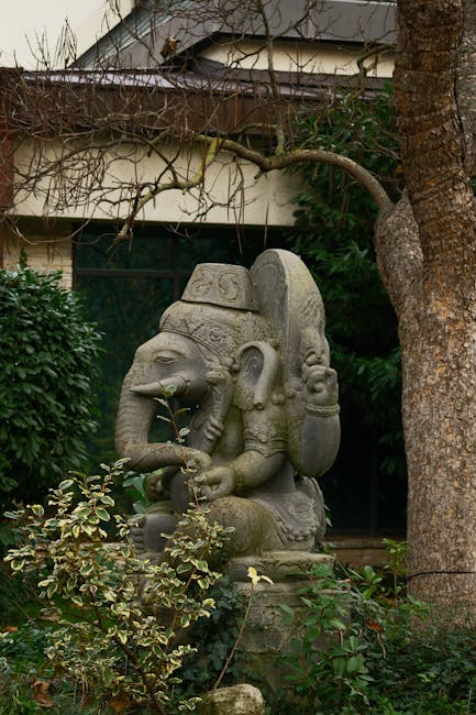 A modern urban garden situated alongside a sleek, curved glass building reflecting surrounding trees and sky. The garden features a landscaped section with lush green foliage, small trees with texture